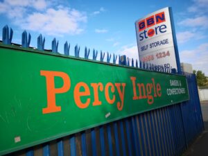 Large green hoarding with the words: Percy Ingle Bakers & Confectioners in orange and white writing on blue metal railings.