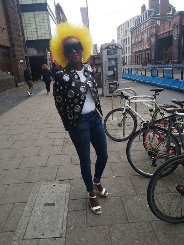 A woman with a bright yellow afro, big sunglasses and a leather jacket smiles into a camera. In the background a couple walks along next to a man who looks back towards the woman.