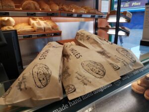 Three brown paper bags with pictures of bread and words reading 'Ancient Grains Sour Dough on top of a counter with loaves of bread in the background.