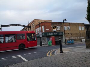Red single-decker bus with 'Go' logo rides under and Arch reading 'ROMAN ROAD' with Tower Hamlets logo. Closed Ingles bakery shopfront with 'To Let' sign above. On the wall is written: 'MAKE THE RICH PAY FOR COVID 19'.