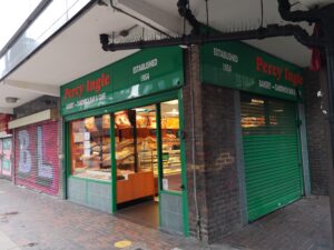 A shop front reading Percy Ingle in orange on green backdrop. White writing reads: 'Bakery - Sandwich Bar & Cafe' and 'Established 1954'