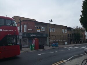 Red double decker bus with 'Stagecoach' logo rides under and Arch reading 'ROMAN ROAD' with Tower Hamlets logo. Closed Ingles bakery shopfront with 'To Let' sign above. On the wall is written: 'MAKE THE RICH PAY FOR COVID 19'.