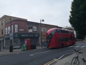 Red double decker bus with 'Stagecoach' logo with number 8 on its back drives down the road beyond 'ROMAN ROAD' with Tower Hamlets logo. Closed Ingles bakery shopfront with 'To Let' sign above. bus covers a logo written on wall with the word : 'MAKE' visible.