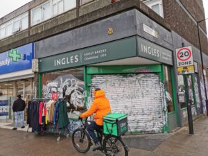 Man wearing bright orange jacket rides a bike with Uber Eats logo past a shut Ingles Bakery. In front of the store is a rack of clothes with a '£2' sign.
