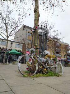 A cycle painted white is tied to the railings around a tree. There is festive tinsel on the bike and on the railings.