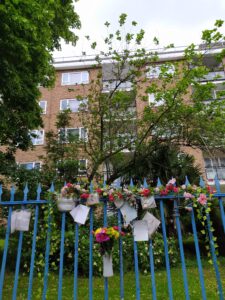 A bunch of flowers in a white vase and handwritten notes are attached to blue railings with a spiky top that are in front of a block of flats. There are artificial flowers and ivy near the top of the railings.