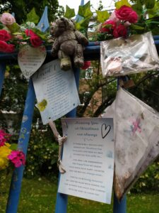 A note attached to blue railings says 'Missing you at Christmas and always with a heart sign and the text of a letter. A weather damaged teddy and artificial flowers are at the top of the railings.